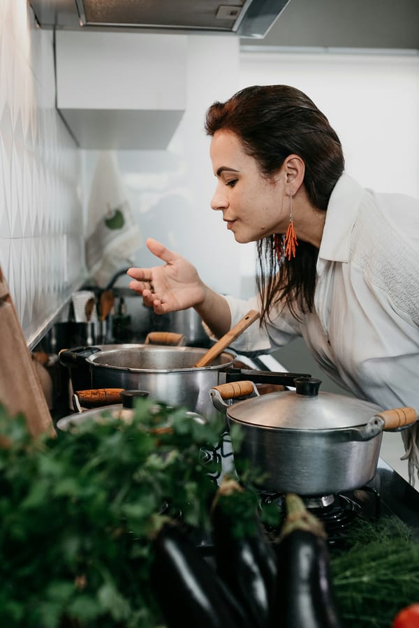 A woman leaning over two pans on her stove, alone in her kitchen, herbs and aubergines in the foreground