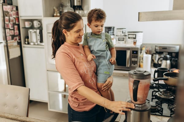 A mother holding a toddler on one hip while running a blender one-handed beside the stove