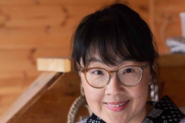 Shihoko Ura of Chopstick Chronicles smiling in her kitchen, wearing round glasses, against a wooden wall